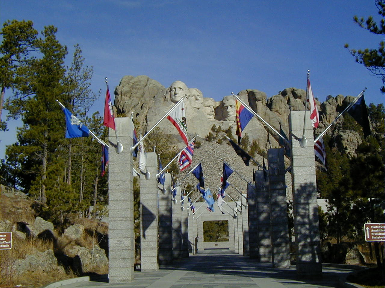 A walkway lined with stone pillars and flags leads to Mount Rushmore, where four U.S. presidents’ faces are carved into the rocky mountain, surrounded by trees under a clear blue sky—an iconic spot to watch the Mount Rushmore fireworks 2026.