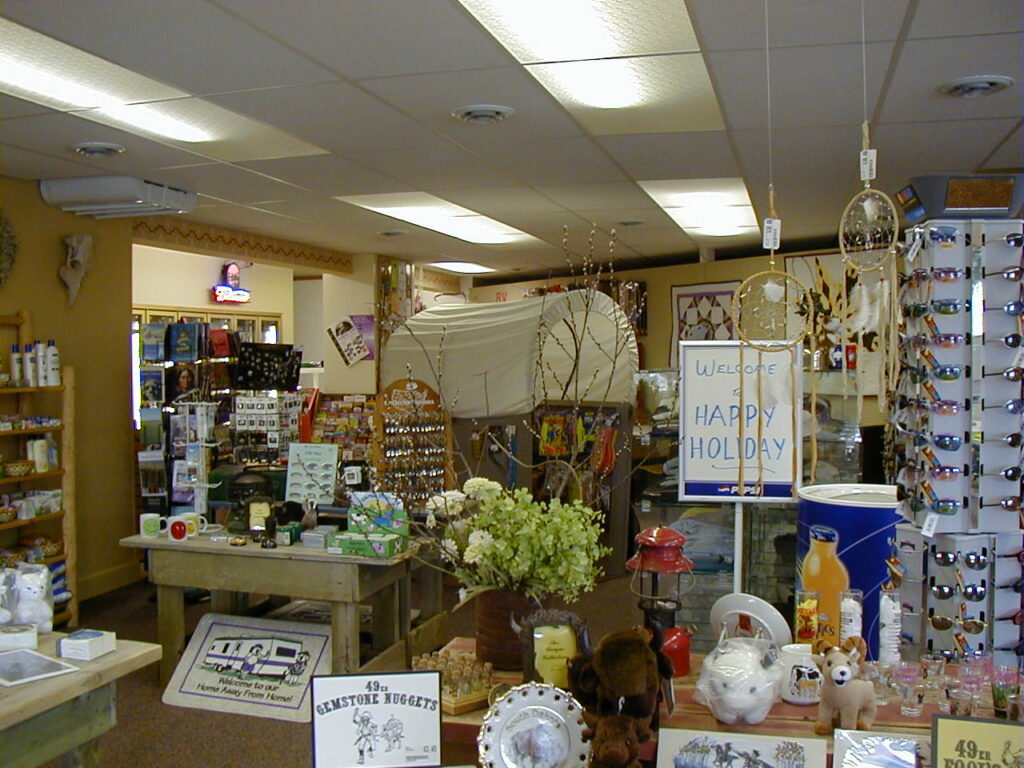 A gift shop interior filled with various items including souvenirs, sunglasses, dreamcatchers, plush toys, and flowers. Shelves and tables display assorted goods perfect for celebrating Mount Rushmore fireworks 2026. A sign reads "Welcome Happy Holiday.