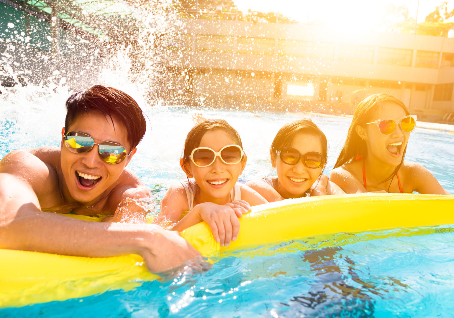 Four people wearing sunglasses smile and lean on a yellow inflatable in a swimming pool, with water splashing behind them and sunlight shining in the background.