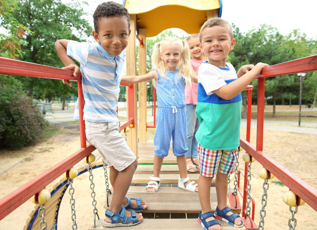 Four smiling children stand on a wooden bridge at a playground, holding onto red railings. Trees and park grounds are visible in the background, creating a cheerful and playful outdoor scene.
