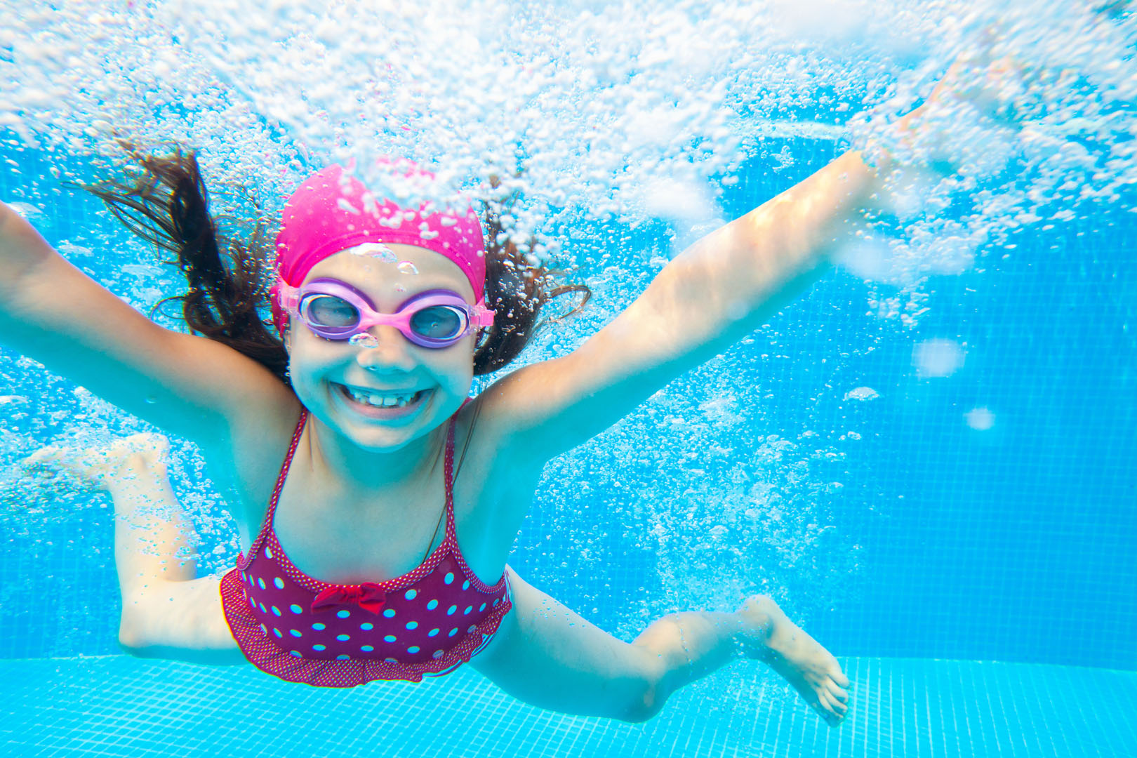 A young girl in a pink swim cap, goggles, and a red polka-dot swimsuit swims underwater in a pool, smiling with arms outstretched and bubbles surrounding her.