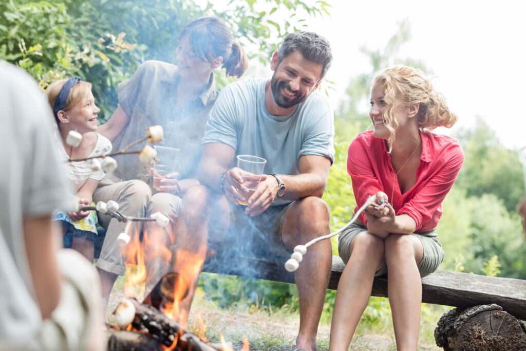 A group of people sit by a campfire outdoors, roasting marshmallows and smiling. Trees and greenery surround them, and the atmosphere is cheerful and relaxed.