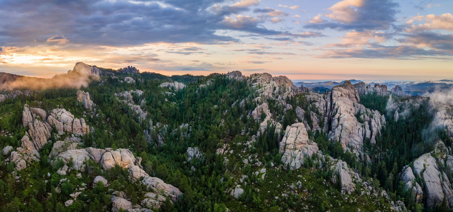 A panoramic view of rugged, rocky hills covered with dense green pine trees under a partly cloudy sky at sunrise or sunset, with light mist and distant mountains on the horizon.