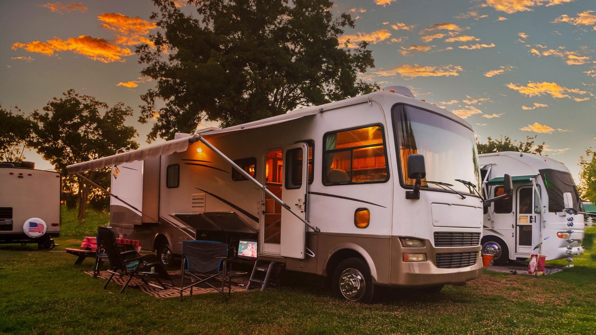 A large RV with an extended awning is parked on grass at sunset, surrounded by chairs and outdoor gear, with another RV nearby and trees silhouetted against a colorful sky.