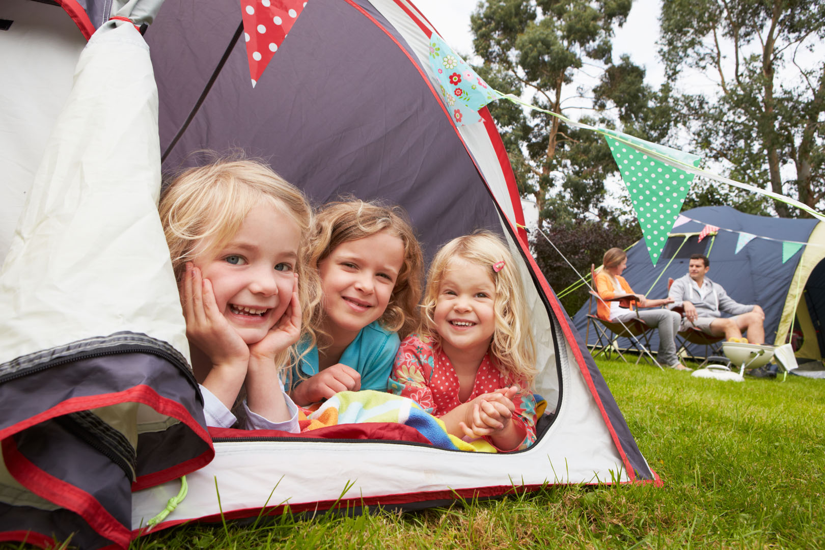 Three smiling children lie inside a tent at a campsite, with colorful bunting outside. In the background, two adults sit by another tent, enjoying the outdoor setting. Trees and grass are visible around them.