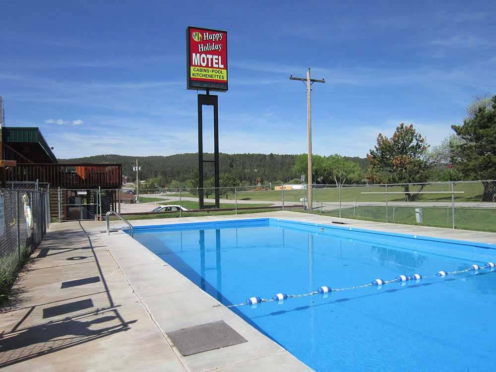 A clear outdoor swimming pool is surrounded by a concrete deck and chain-link fence, with a “Happy Holiday Motel” sign visible in the background and green hills under a blue sky.