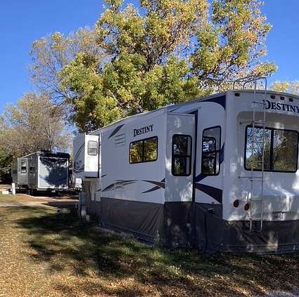 Two white RVs are parked beneath leafy trees in a sunlit campground. The nearest RV has the word “DESTINY” on its rear, and both sit on grassy ground with scattered shade and blue sky above.