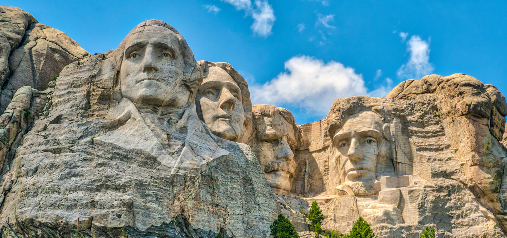 Mount Rushmore National Memorial featuring the carved faces of four U.S. presidents on a granite cliff, set against a blue sky with clouds and surrounded by rocky landscape and some greenery.