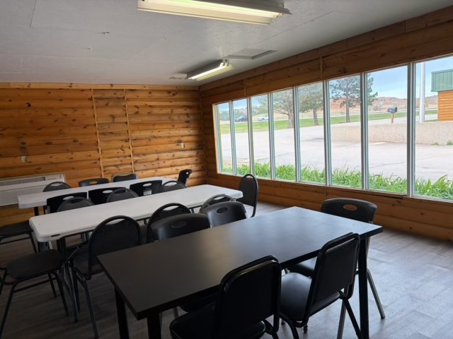 A room at Amenities Happy Holiday RV Resort features wooden log walls, large windows, and two tables—one white, one black—surrounded by black chairs. Outside are trees and a parking lot. Fluorescent lights illuminate the space.